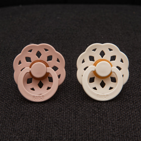 Close-up of two pacifiers in blush pink and ivory from the Lola Pacifier Set, displayed on a textured black surface.