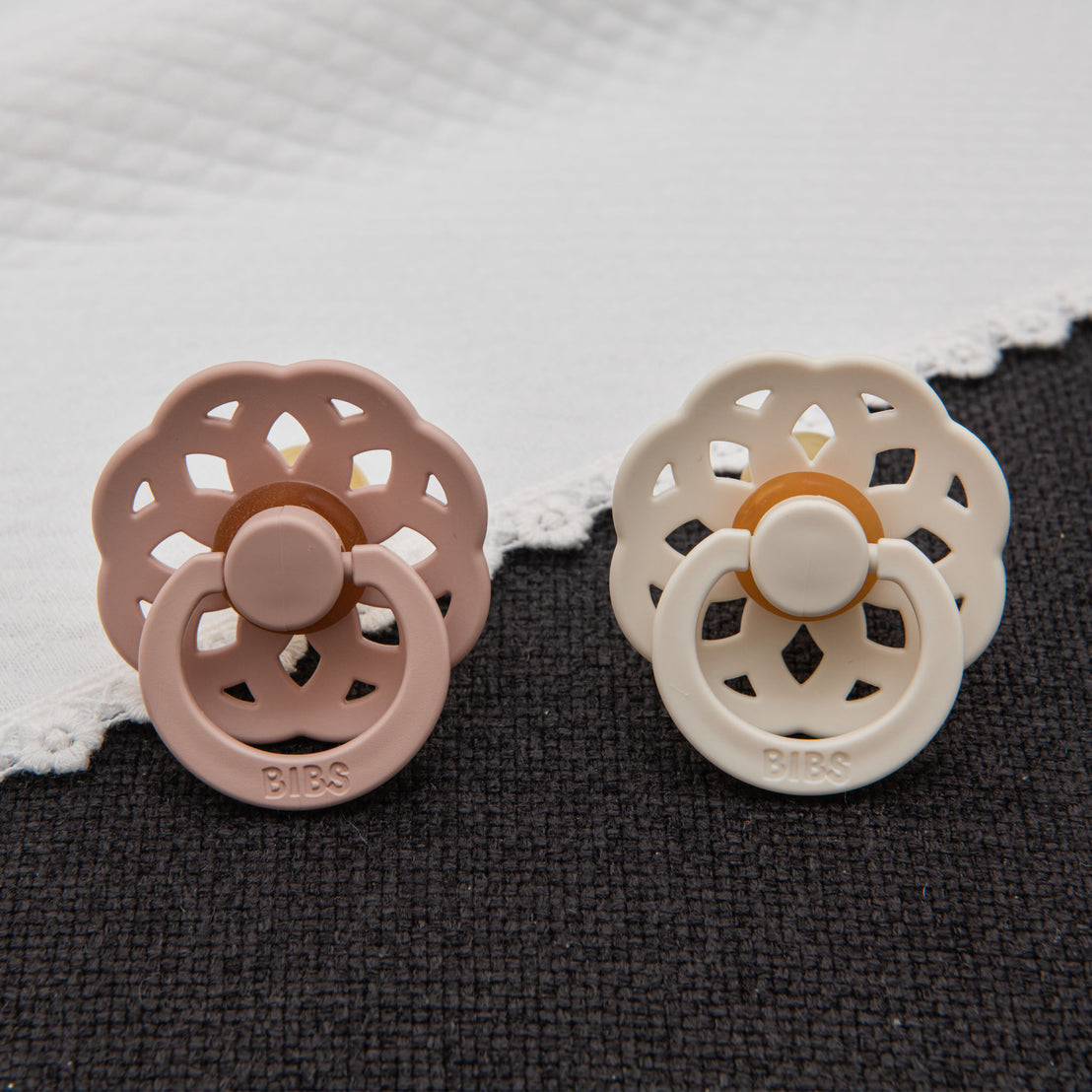 Blush pink and ivory pacifiers from the Lola Pacifier Set displayed against a dark textured fabric, with a white quilted blanket in the background.