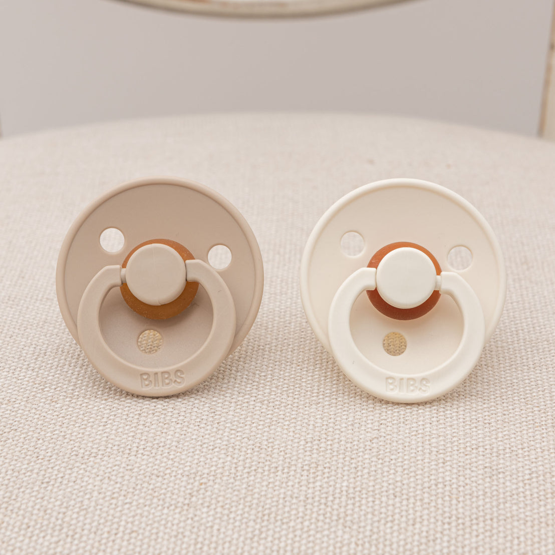 Two upscale natural-colored Braden Pacifier Sets placed on a textured fabric surface, perfect as a baby gift.
