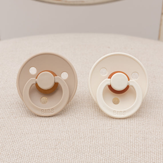 Two upscale natural-colored Braden Pacifier Sets placed on a textured fabric surface, perfect as a baby gift.