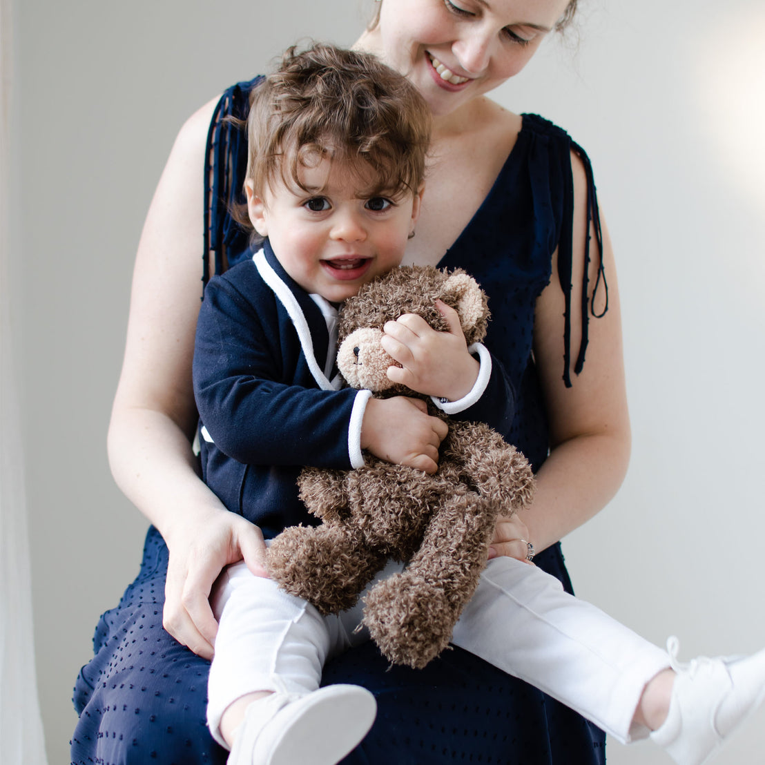 Smiling baby boy sitting on his mother's lap. He is wearing the Elliott 3-Piece Suit, including the jacket, pants, and onesie. He is also holding the Cubby Bear.