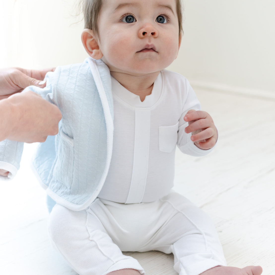 Baby boy sitting on the ground and wearing the Logan 3-Piece Suit, including the jacket, pants and onesie. Jacket made from blue textured cotton with a white French terry cotton trim.