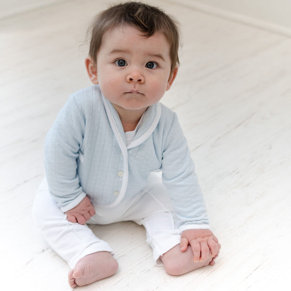 Baby boy sitting on the ground and wearing the Logan 3-Piece Suit, including the jacket, pants and onesie.  Jacket made from blue textured cotton with a white French terry cotton trim.