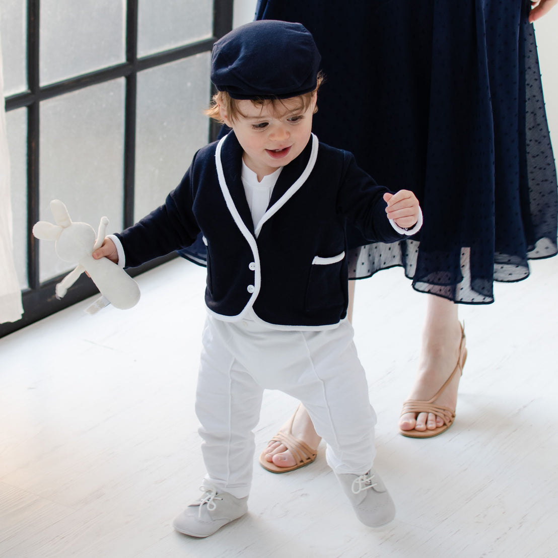 Smiling baby boy walking with his mother behind him. He is wearing the Elliott 3-Piece Suit, including jacket, pants, and onesie (and matching newsboy cap).