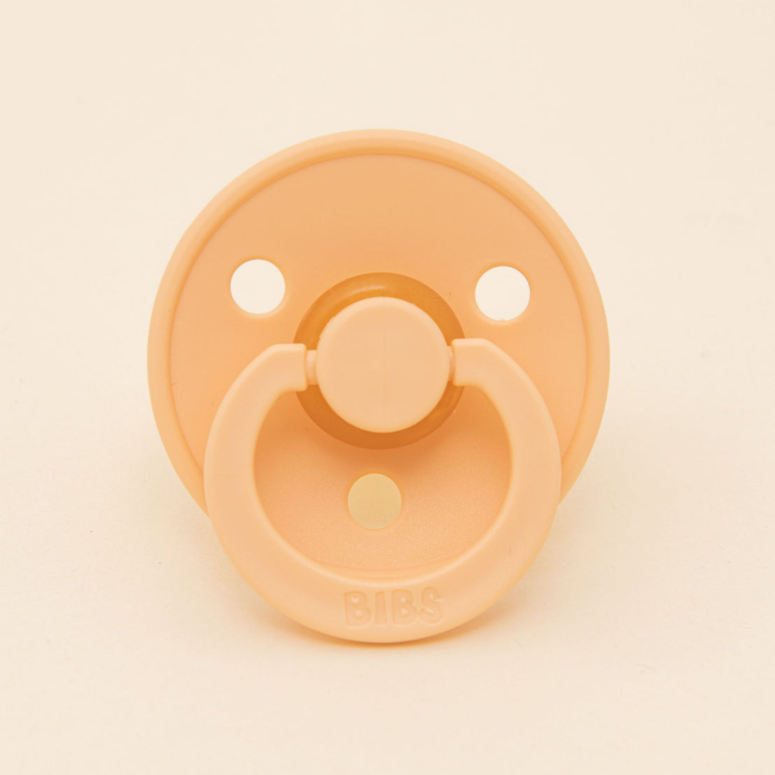 A Bibs pacifier in Peach Sunset with a round shield and two air holes, prominently featuring the brand name "bibs" on the button.