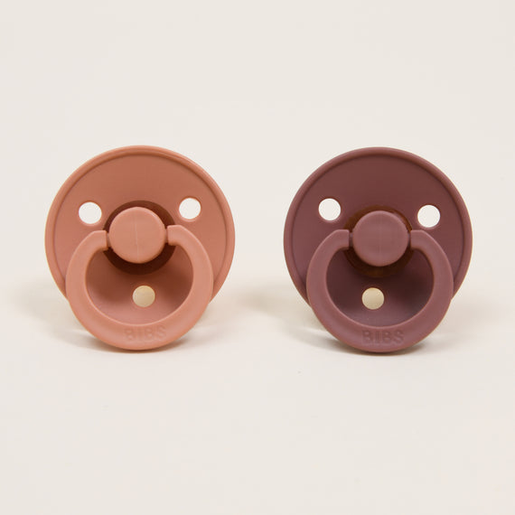Two Bibs Pacifier Sets in Peach Sunset and Woodchuck colors, displayed against a light background. Each pacifier has a round design and a button handle.