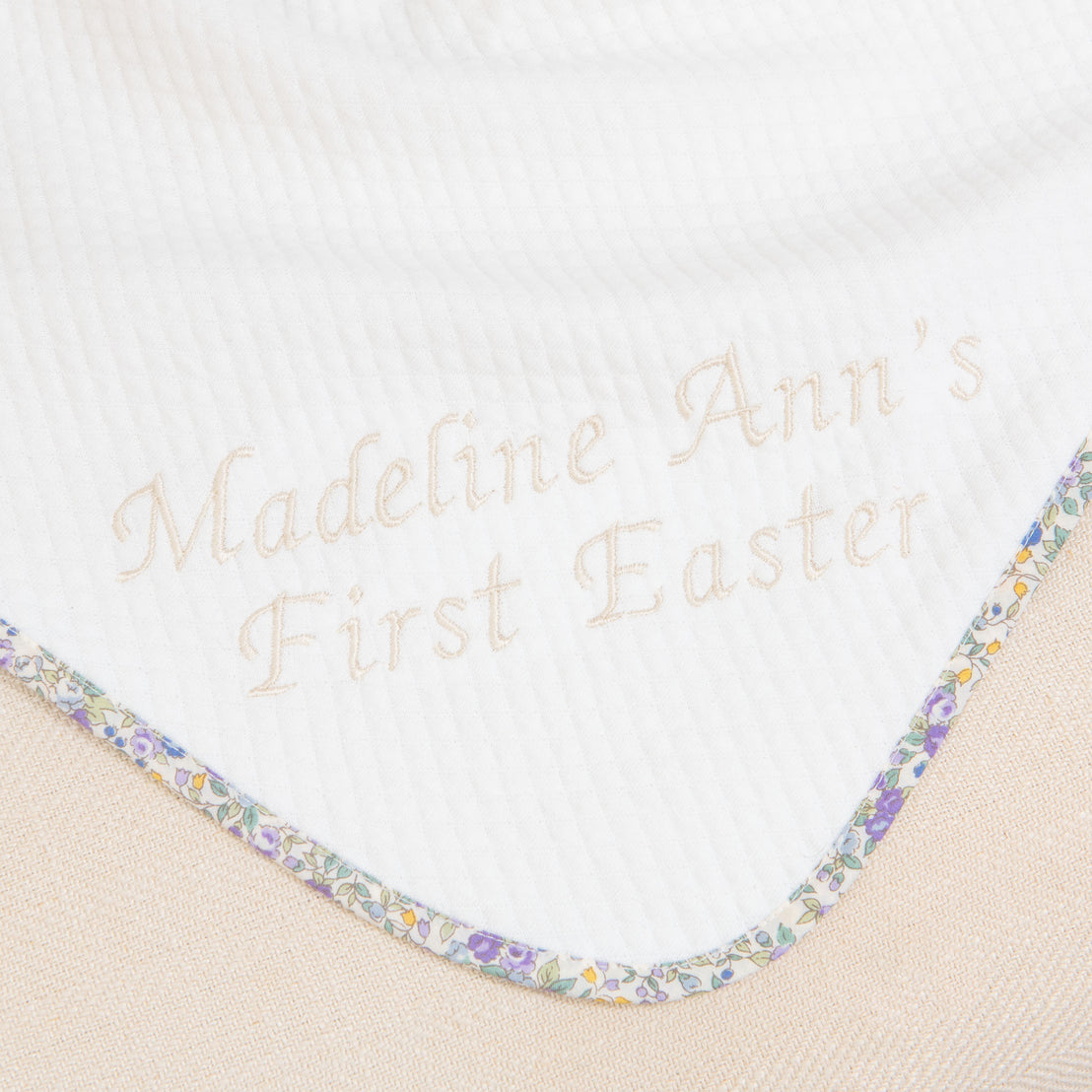 Close-up of a Petite Fleur Gift Set - Save 10% embroidered with the words "Madeline Ann's first Easter" surrounded by a floral border on beige fabric.