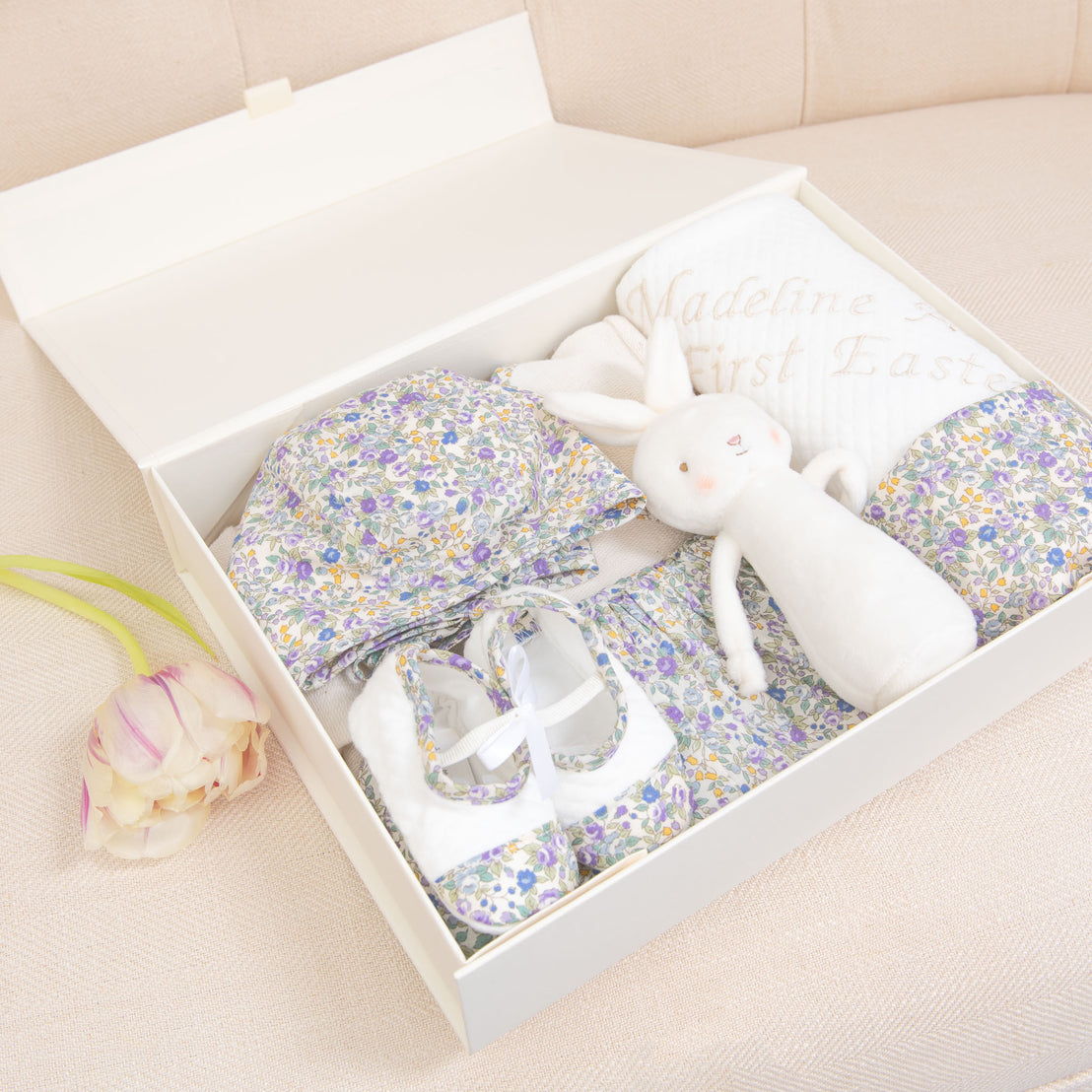 An upscale boutique Petite Fleur Gift Set containing baby items: a vintage-inspired floral dress, matching shoes, and a white bunny plush toy. A ribbon and custom tag read "Madeline's First Easter". Save 10%