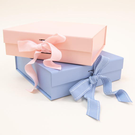 Two sophisticated "Gift Wrap | Small Gift Box with Bow" packages are artistically arranged against a soft backdrop. A pink box adorned with a delicate pale pink ribbon pairs beautifully with a blue box, which is accented by a blue ribbon featuring white stitching. Ideal for any occasion, these charming boxes embody the elegance of Baby Beau & Belle.