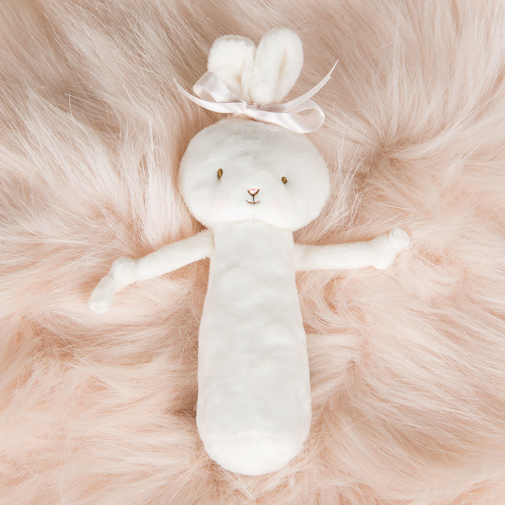 The Emma Bunny Rattle, a plush white baby rattle with a bow, is set against a soft pink fluffy background, making it ideal for an upscale boutique baptism gift.