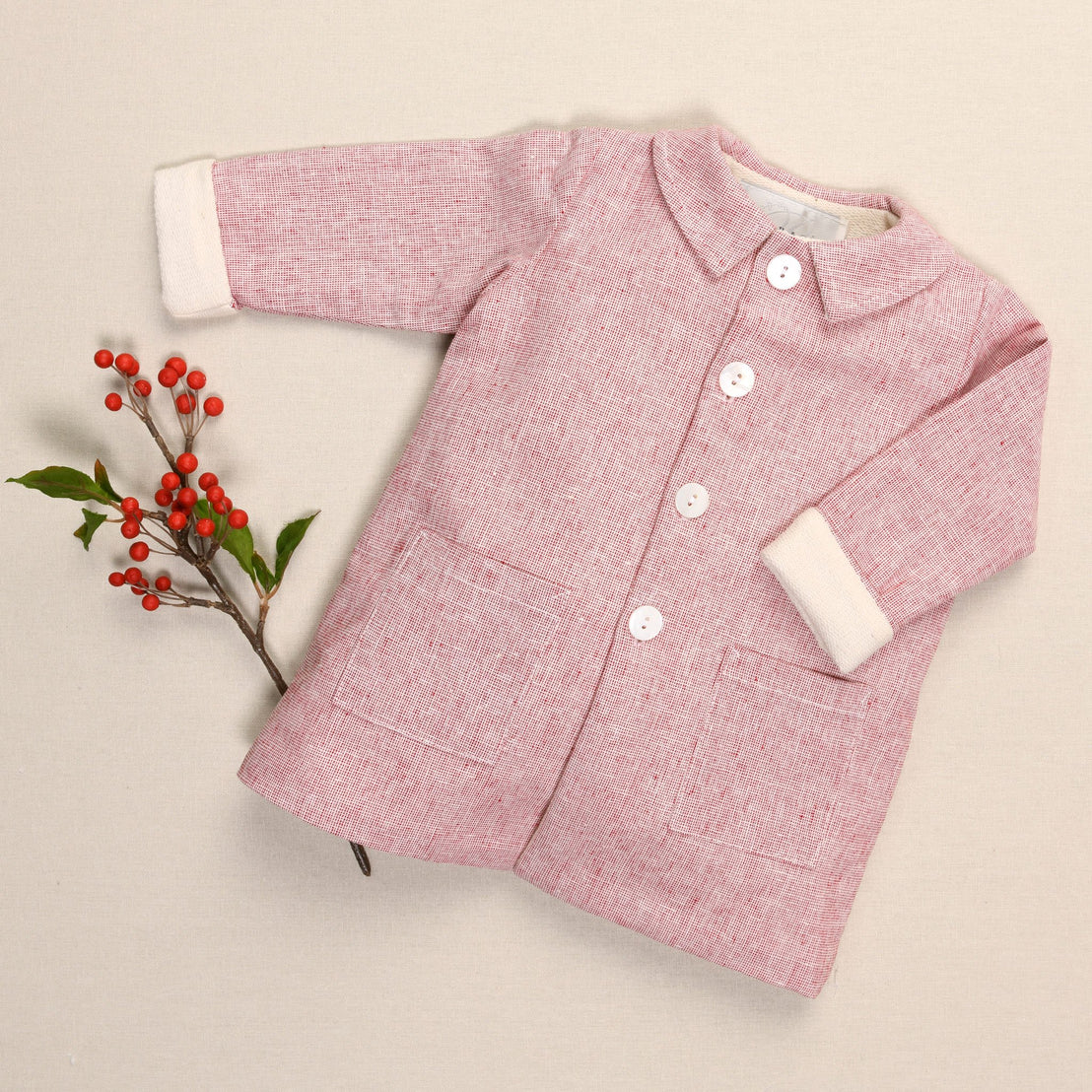 A handmade red linen trench coat for children, adorned with white buttons and two pockets, rests gently against a beige background, accented by a sprig of red berries and green leaves to its left.