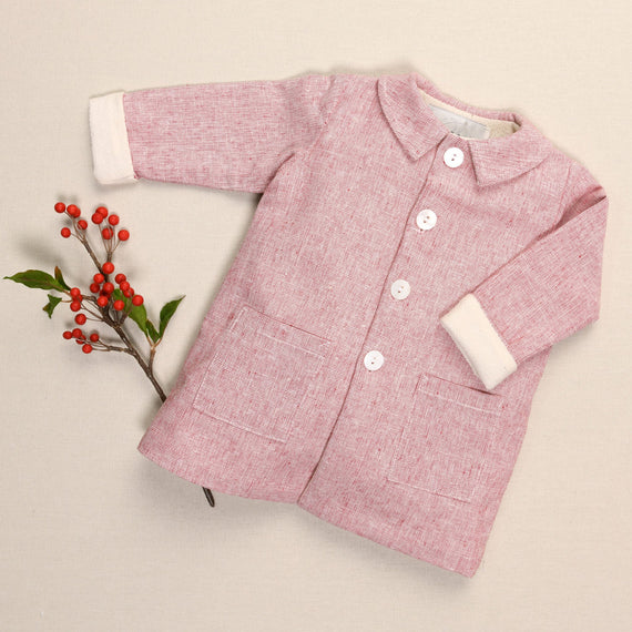 A handmade red linen trench coat for children, adorned with white buttons and two pockets, rests gently against a beige background, accented by a sprig of red berries and green leaves to its left.