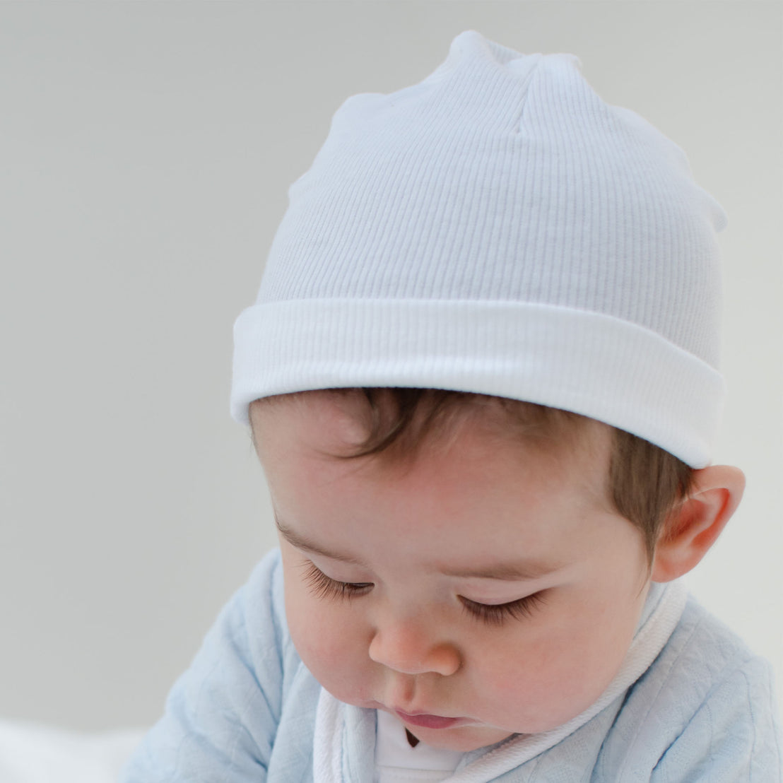 A baby is wearing a Natural Ivory Ribbed Pima Beanie and a light blue outfit, gazing downward. The bright, simple background highlights the baby's serene and attentive expression.