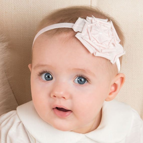 A close-up of a baby with bright blue eyes wearing an Emma Linen Rose Headband, looking curiously at the camera.