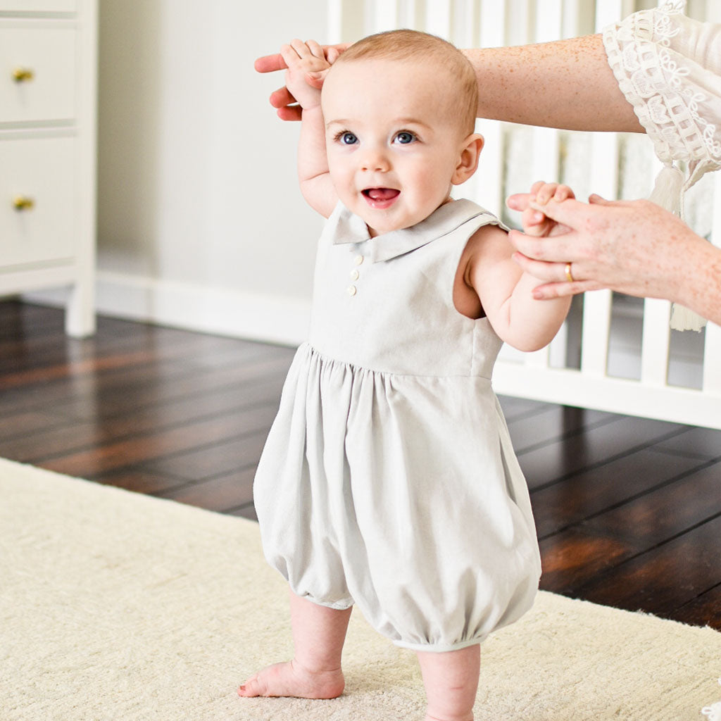 Baby boy walking with the help of his mother. He is wearing the Grayson Linen Romper, a bubble romper made from a soft linen cotton blend in grey and features a pointed collar and button detail on the front.