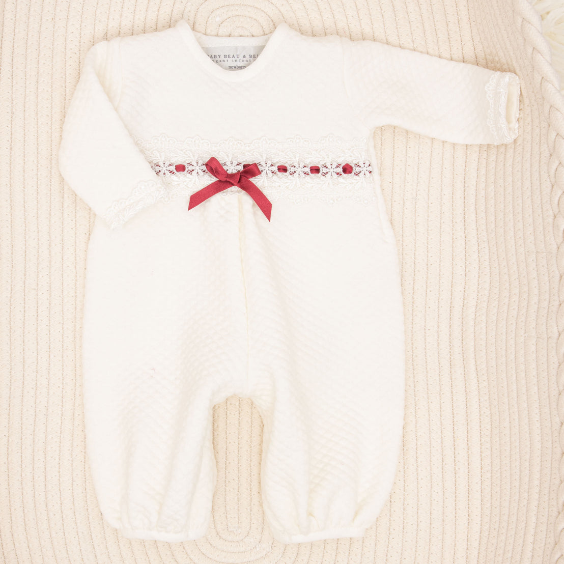 The Clara Gift Set, featuring a white baby romper adorned with lace details and a red ribbon bow across the chest, handcrafted from quilted cotton for maximum comfort, arranged on a textured beige surface, is currently available with a 10% discount.