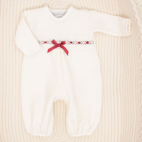Introducing the Clara Holiday Romper: a cozy, white newborn jumpsuit featuring long sleeves and a textured pattern. It is beautifully embellished with delicate ivory lace detailing across the chest and adorned with a charming red ribbon bow. The romper is elegantly presented on a soft, cream-colored surface.