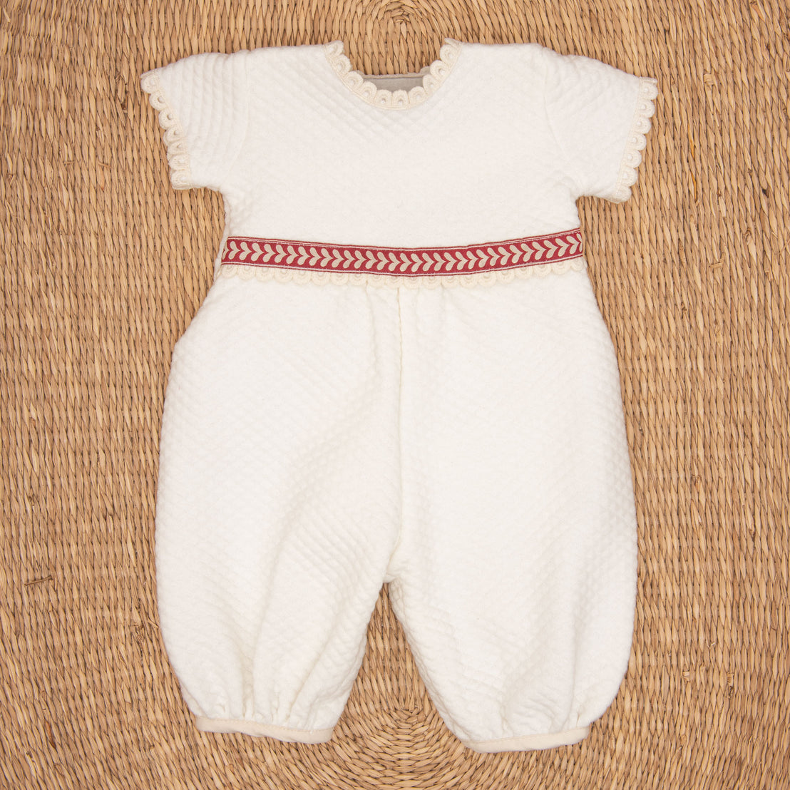 The Valentina Romper is a white infant outfit with short sleeves and textured fabric, featuring delicate lace edging. Ideal for a newborn Christmas ensemble, it has a red and white herringbone patterned band at the waist. The romper is displayed flat against a woven straw mat background.