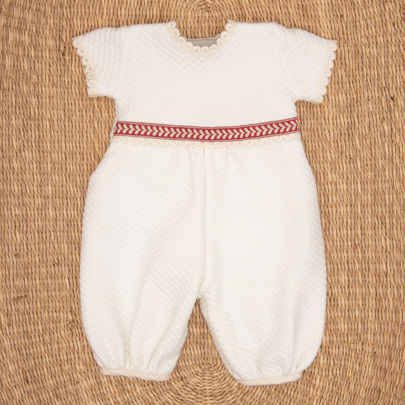 The Valentina Romper is a white infant outfit with short sleeves and textured fabric, featuring delicate lace edging. Ideal for a newborn Christmas ensemble, it has a red and white herringbone patterned band at the waist. The romper is displayed flat against a woven straw mat background.