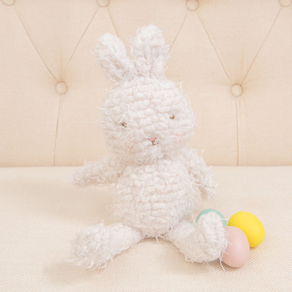 The Shaggy Hoppy Bunny, with its long ears and plush, textured fur that feels just like a dream, sits on a cream-colored couch. Beside the bunny are two small, colorful plastic eggs, one yellow and one pink—ideal for a baby shower gift.