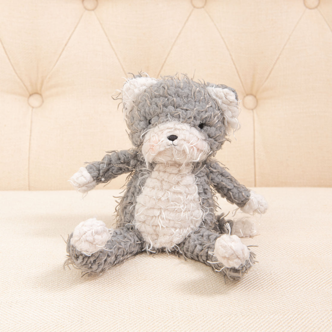 A plush Shaggy Purrty Kitty toy with soft grey and white fur rests on a beige, tufted fabric background. Perfect as a baby shower gift, the cuddly cat has a relaxed pose and a gently worn appearance.