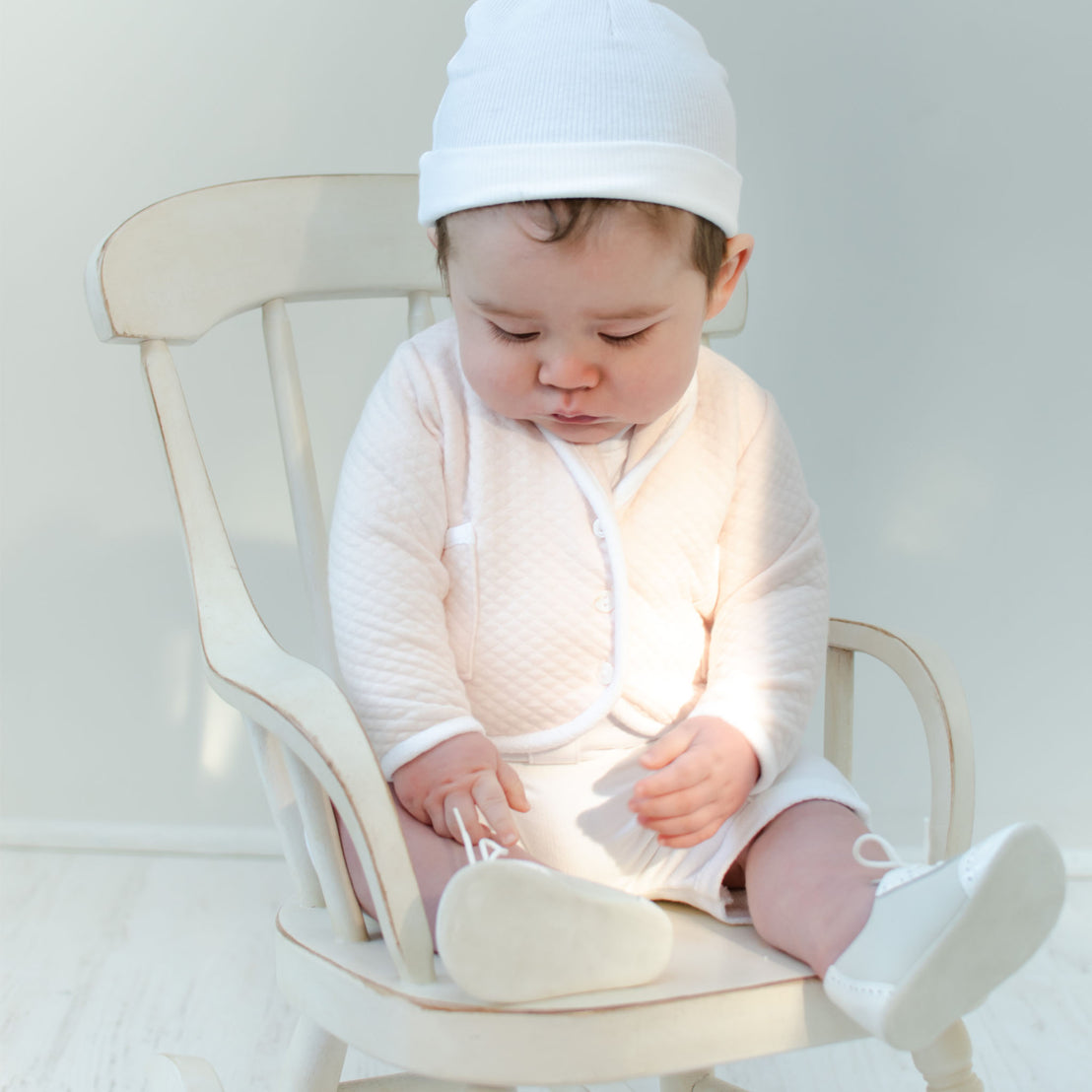 A baby, dressed in the Asher Blush 3-Piece Shorts Suit, sits on a wooden rocking chair, gazing down at their feet. The setting is bright and softly lit, creating a peaceful and serene atmosphere.