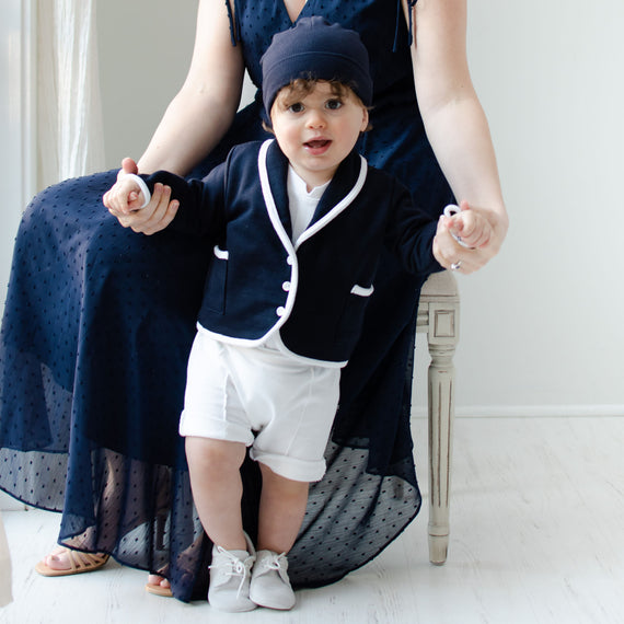 Baby boy holding on to his mother's hands. He is wearing the Elliott 3-Piece Shorts Suit, including the jacket, shorts, and onesie (and matching Navy Ribbed Pima Beanie).