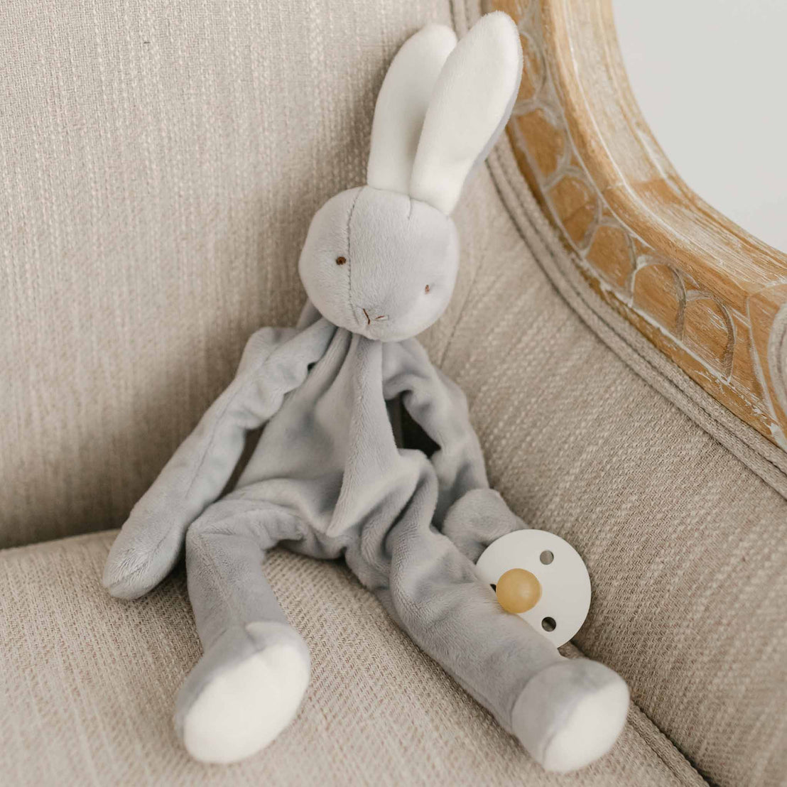 A flat lay photo of the Miles Silly Bunny Pacifier Holder. It is a grey bunny stuffed animal that can hold onto a pacifier