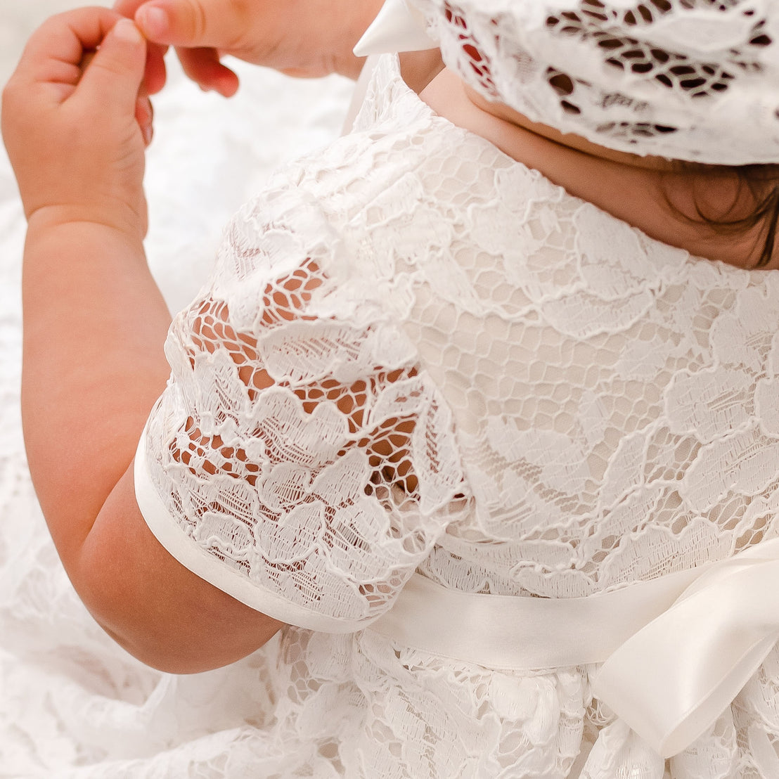 A close-up of a baby dressed in the Rose Christening Gown & Bonnet for baptism, holding an adult's finger. The baby also wears a lace bonnet.