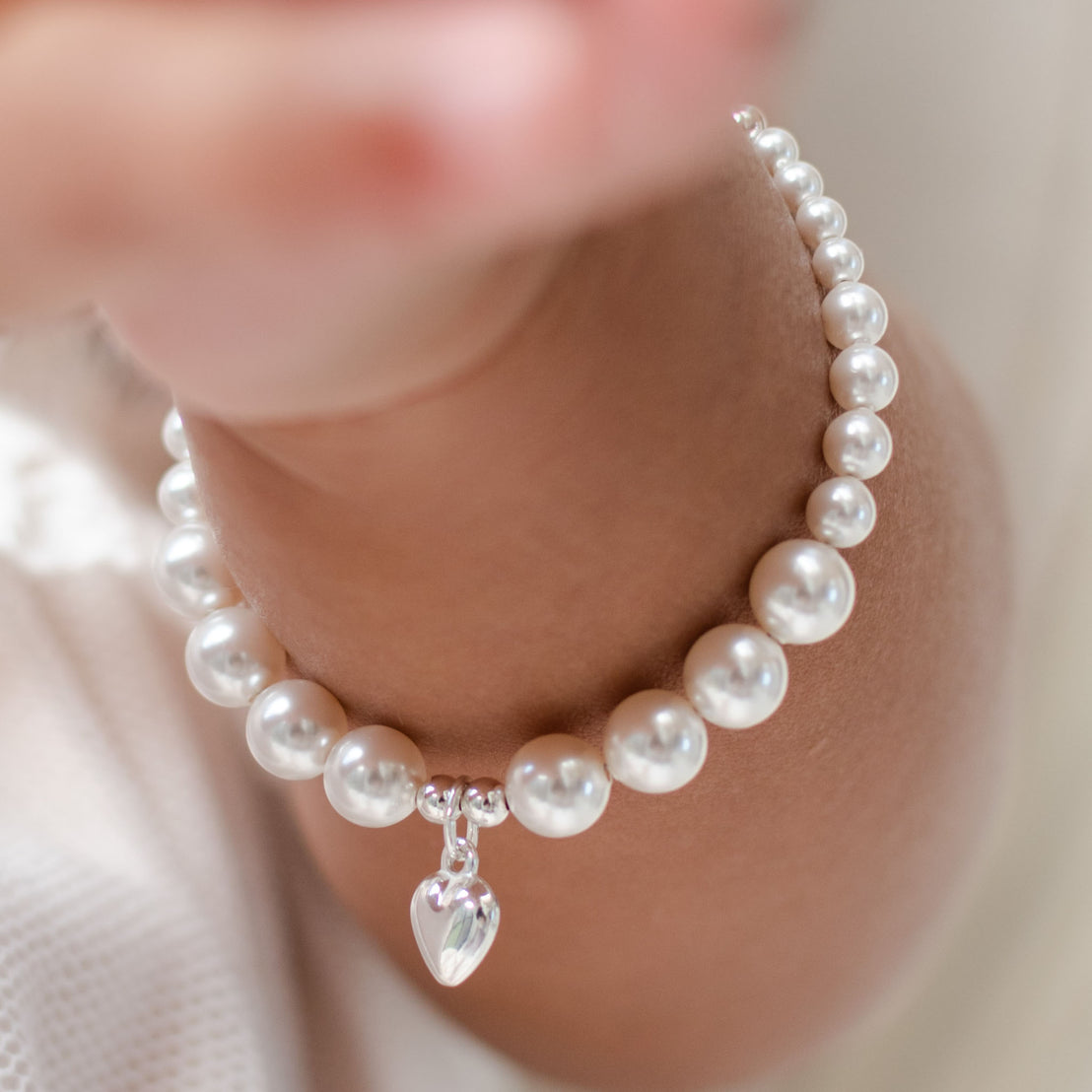 Close-up of the White Luster Pearl Bracelet with a silver heart charm draped on a baby's wrist, highlighting the shiny pearls and delicate charm.