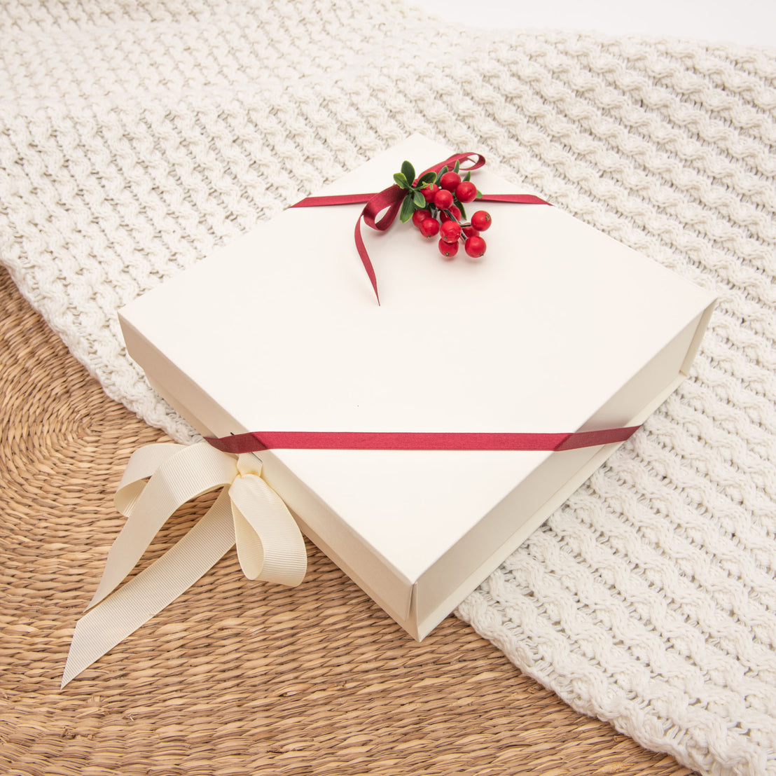 A square Christmas gift wrap tied with a red ribbon, adorned with red berries and green leaves on top, resting on a textured white-knit background.