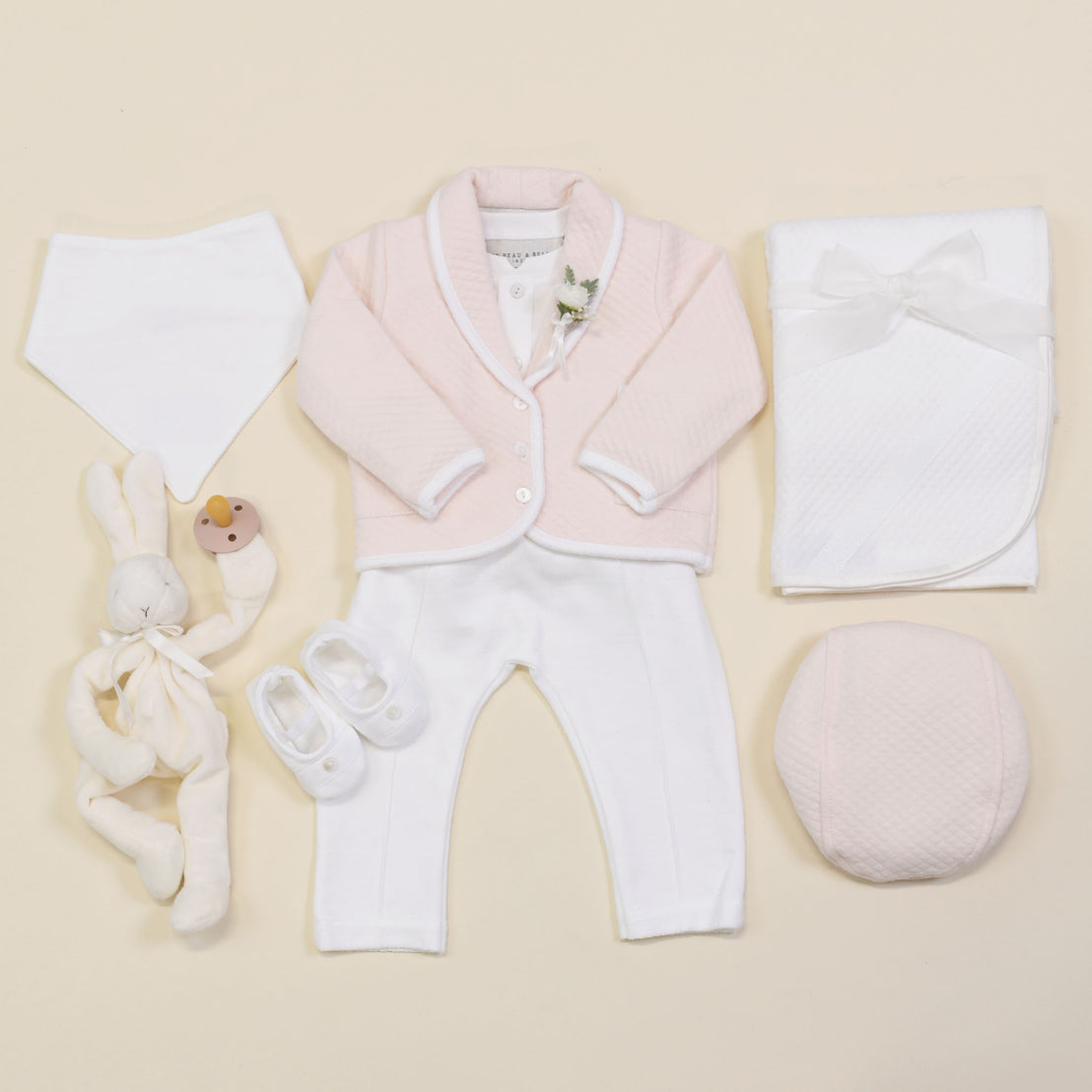 Flat lay of baby items on a neutral background showcasing the Asher Blush 3-Piece Suit, which includes a jacket and pants in pink and white tones, paired with matching shoes. The ensemble is complemented by a white bib. The setup also features a plush rabbit toy with pacifier, cozy quilted cotton blanket, and pink cushion.