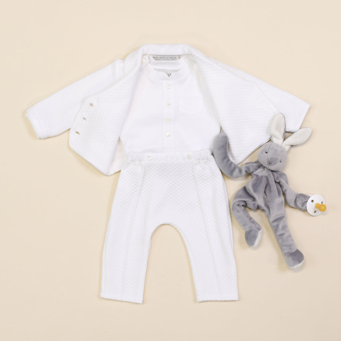 The Elijah Newborn 3-Piece Suit, which includes a white quilted jacket, button-up shirt, and pants, is laid flat. Next to it, a gray stuffed bunny holding a pacifier rests on a light beige background, making it an ideal choice for a baptism outfit.