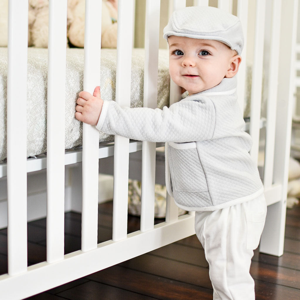 Baby boy standing next to crib wearing his baby boy suit in gray. Part of our Grayson grey baby boy suit collection.