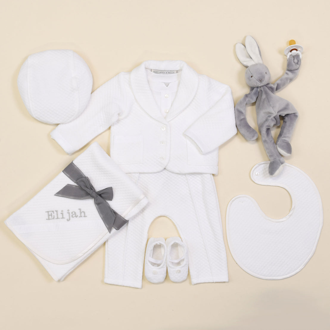 The Elijah Newborn 3-Piece Suit features a white coming home outfit set with a jacket, pants, hat, bib, and booties. Accompanying this charming ensemble is a personalized blanket labeled "Elijah" and a gray rabbit plush toy that holds an orange rattle. Everything is beautifully arranged on a soft beige background.