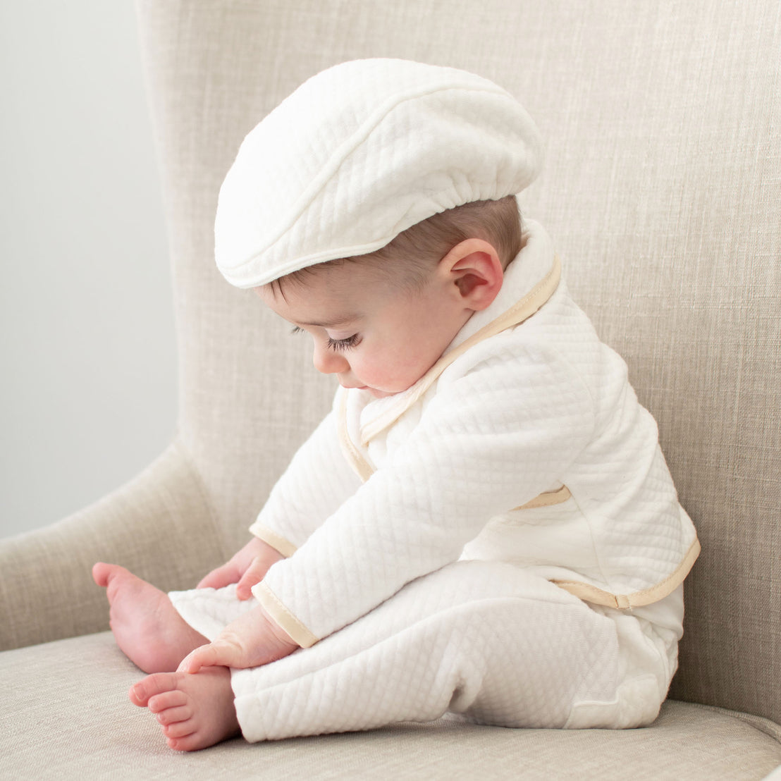Baby boy sitting on a chair. He is wearing the Liam 3-Piece Suit and matching White Quilted Newsboy Cap