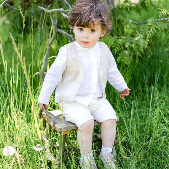 Photo of a baby boy sitting on a wooden chair outside in the forest. He is wearing the Silas Vest Suit that includes the vest, shorts, and onesie