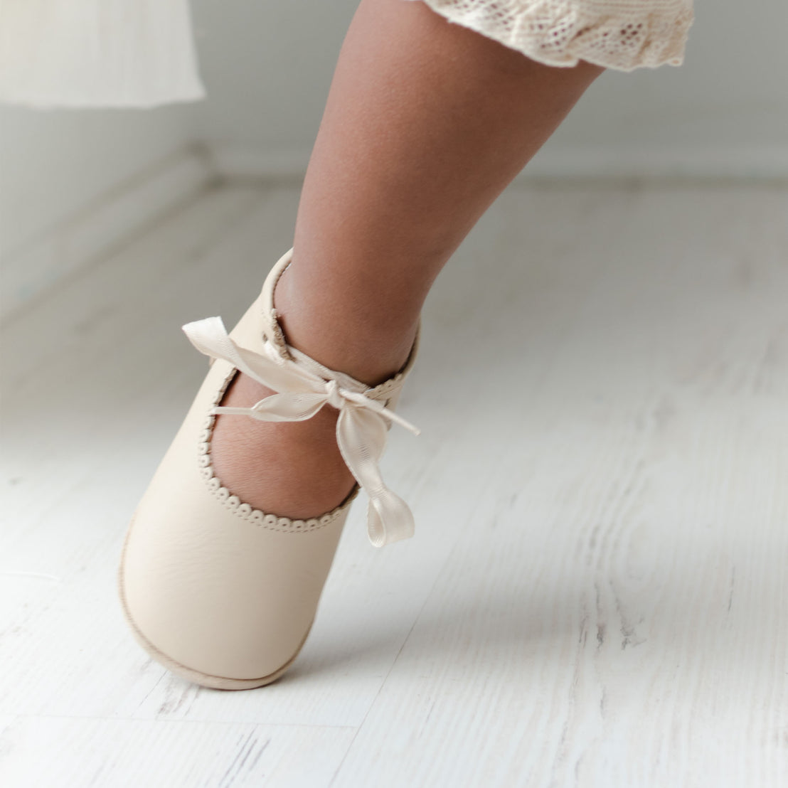 Baby foot wearing the Tan Tie Mary Janes crafted with tan matte leather with beautiful scallop edge detail and complimenting cotton ribbon tie.