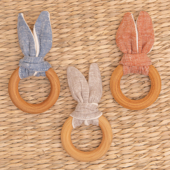 Flat lay photo of all three Silas Wooden Teether Rings, including the colors red, tan, and blue. The teether ring is made from organic maple wood and features a linen and french terry tie