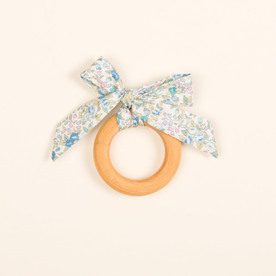 A Petite Fleur Wooden Teether Ring with a delicate floral fabric bow tied around it, presented on a light beige background.