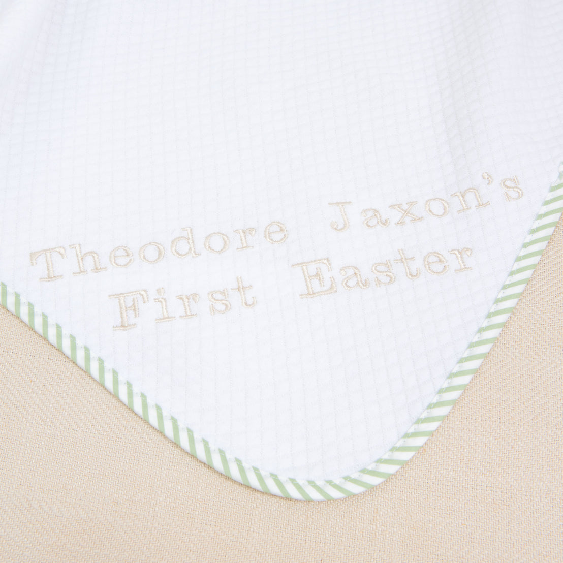 A close-up of the First Easter Personalized Blanket reveals a white fabric with green striped edging, customized with "Theodore Jaxon's First Easter" embroidered in light brown stitches. This personalized keepsake boasts a subtle grid pattern, ideal for celebrating a first Easter.