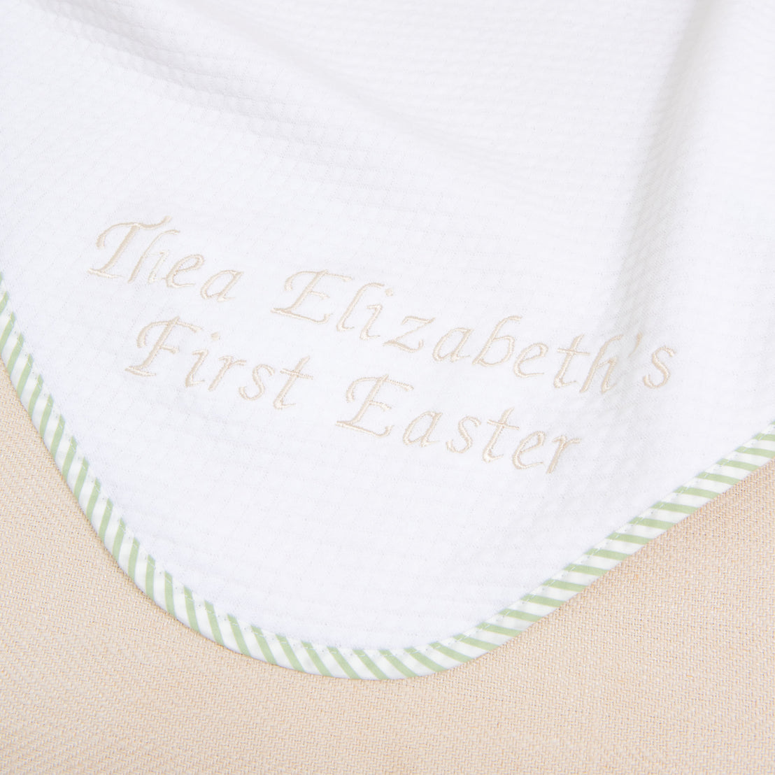 A white blanket featuring green striped edging and custom embroidery that reads "Thea Elizabeth's First Easter" in light brown thread makes it a perfect personalized gift. This is the First Easter Personalized Blanket.
