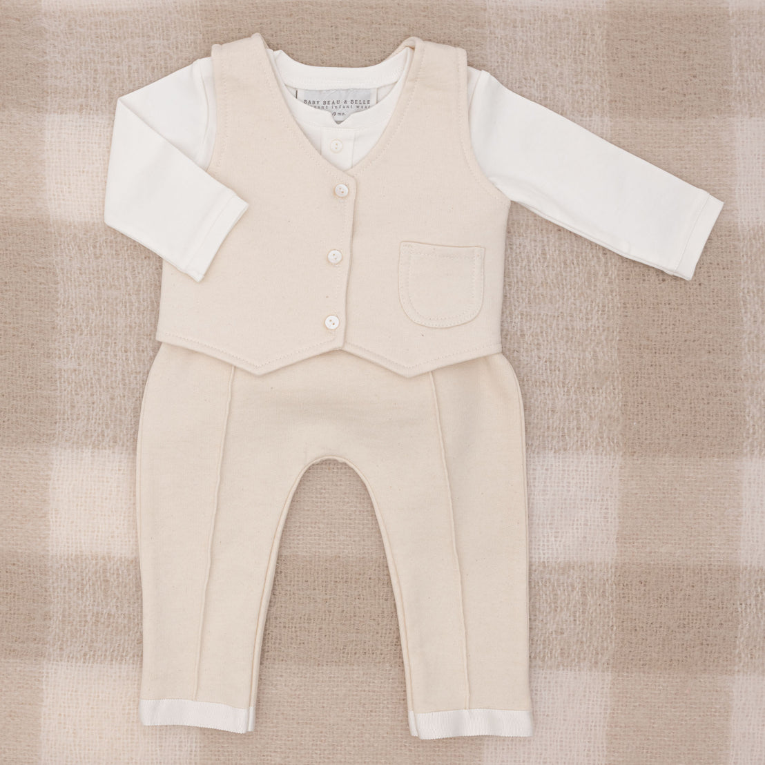 The Braden Vest Suit, a beige baby boy ensemble, includes a sleeveless vest, a white long-sleeve top, and coordinating pants. It is displayed on a textured, beige-checkered fabric background. Made from soft French terry, this suit is ideal for any formal occasion.
