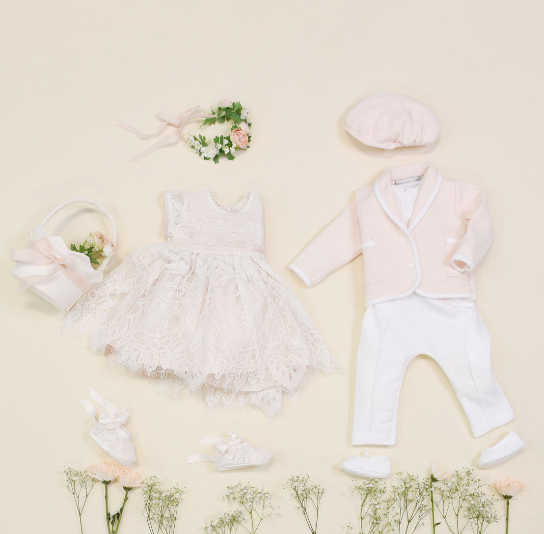 Flat lay of an exquisite Asher Blush 3-Piece Suit on a light background, featuring a lace dress, floral headband, pink jacket, white pants, coordinating hat, shoes, and a basket filled with flowers. The ensemble is arranged amid delicate small white flowers like a quilted cotton dream.