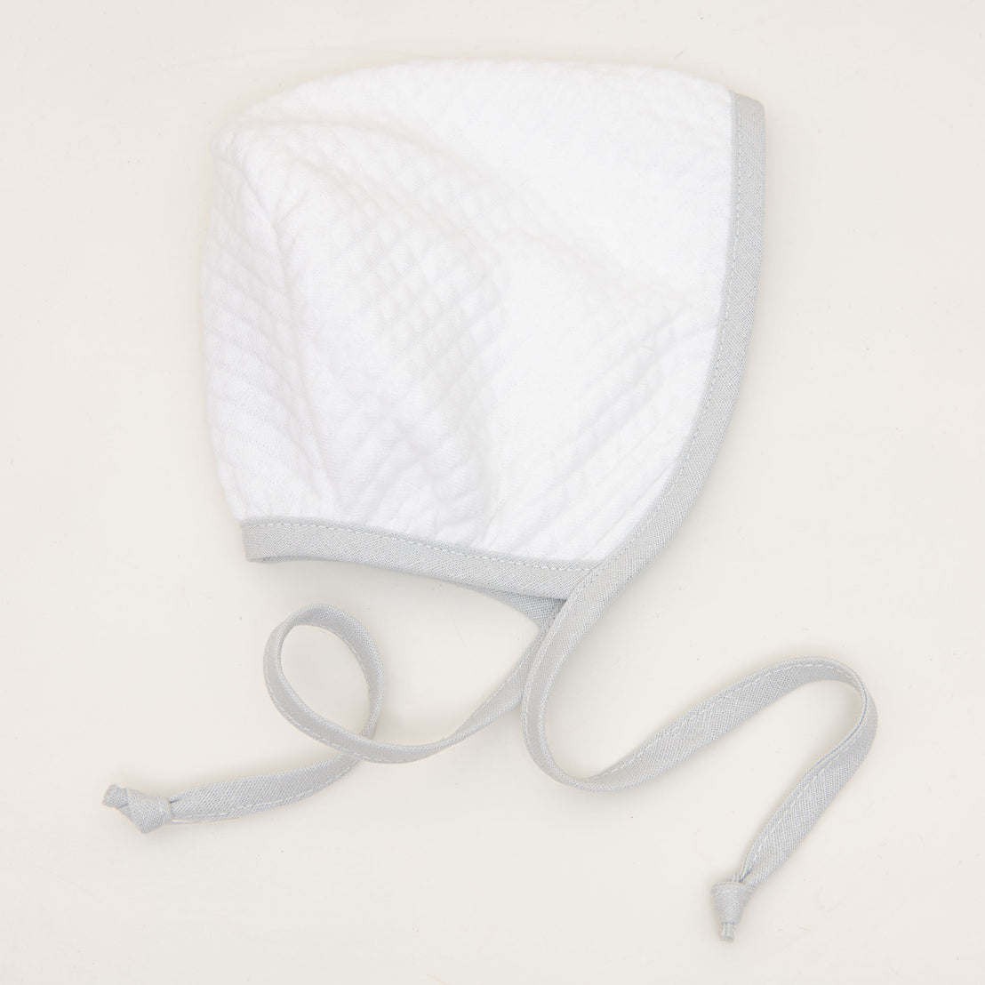 The Grayson White Quilted Bonnet, adorned with light gray edging and long ties, rests on a flat light beige surface. Ideal for a boys' coming home outfit, this classic-style bonnet features a simple and soft texture.