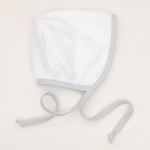 The Grayson White Quilted Bonnet, adorned with light gray edging and long ties, rests on a flat light beige surface. Ideal for a boys' coming home outfit, this classic-style bonnet features a simple and soft texture.