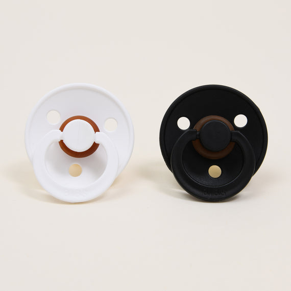 Two Bibs Pacifiers, one white and one black, placed side by side on a light beige background. Both have an upscale look with a button handle and ventilation holes.