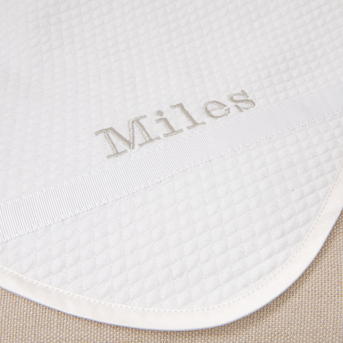 Flat lay photo of the Miles Personalized Blanket on a chair. It is made with quilted cotton and a ribbon detail on the corner. The name "Miles" is embroidered on the corner.