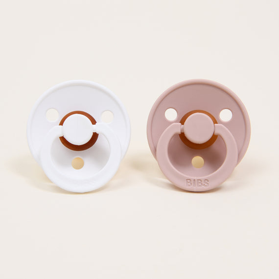 Two upscale Bibs pacifier sets, one white and one blush, placed symmetrically on a light beige background, perfect for a baby gift.