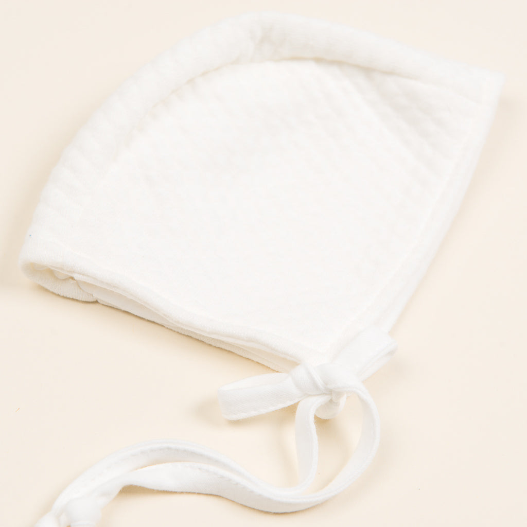 The Ivory Quilt Bonnet, crafted from luxurious Pima cotton, features a soft quilted texture with a tie strap and rests gracefully on a light cream background. Its rounded shape with gently curved edges makes it perfect for any newborn outfit.